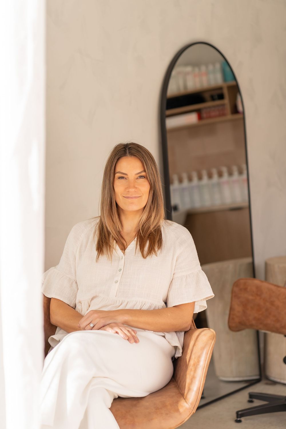 A person in a white linen outfit sits in a brown leather chair at a salon with a mirror visible in the background.