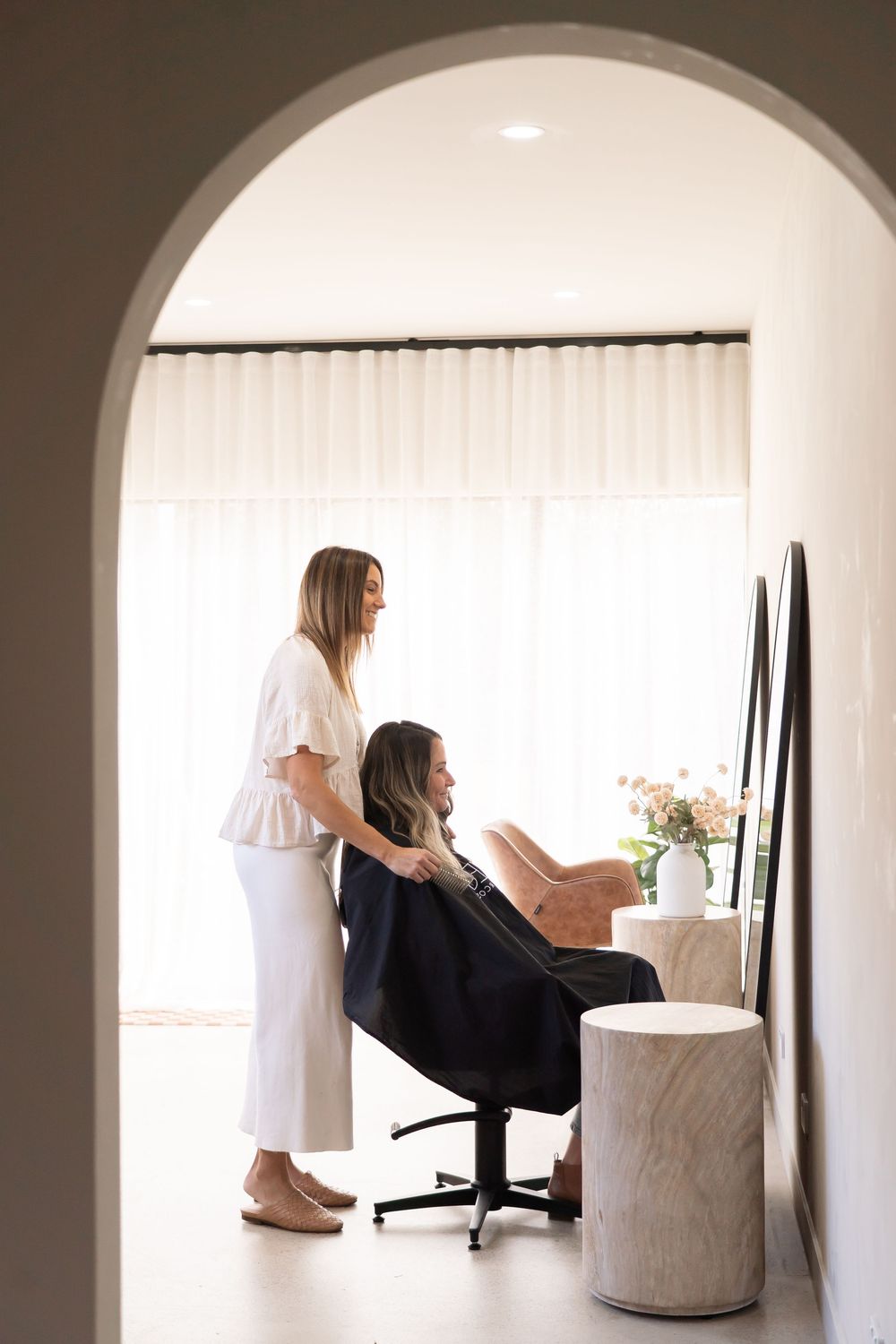 Professional hair stylist working with client in minimalist white salon space with arched doorway.