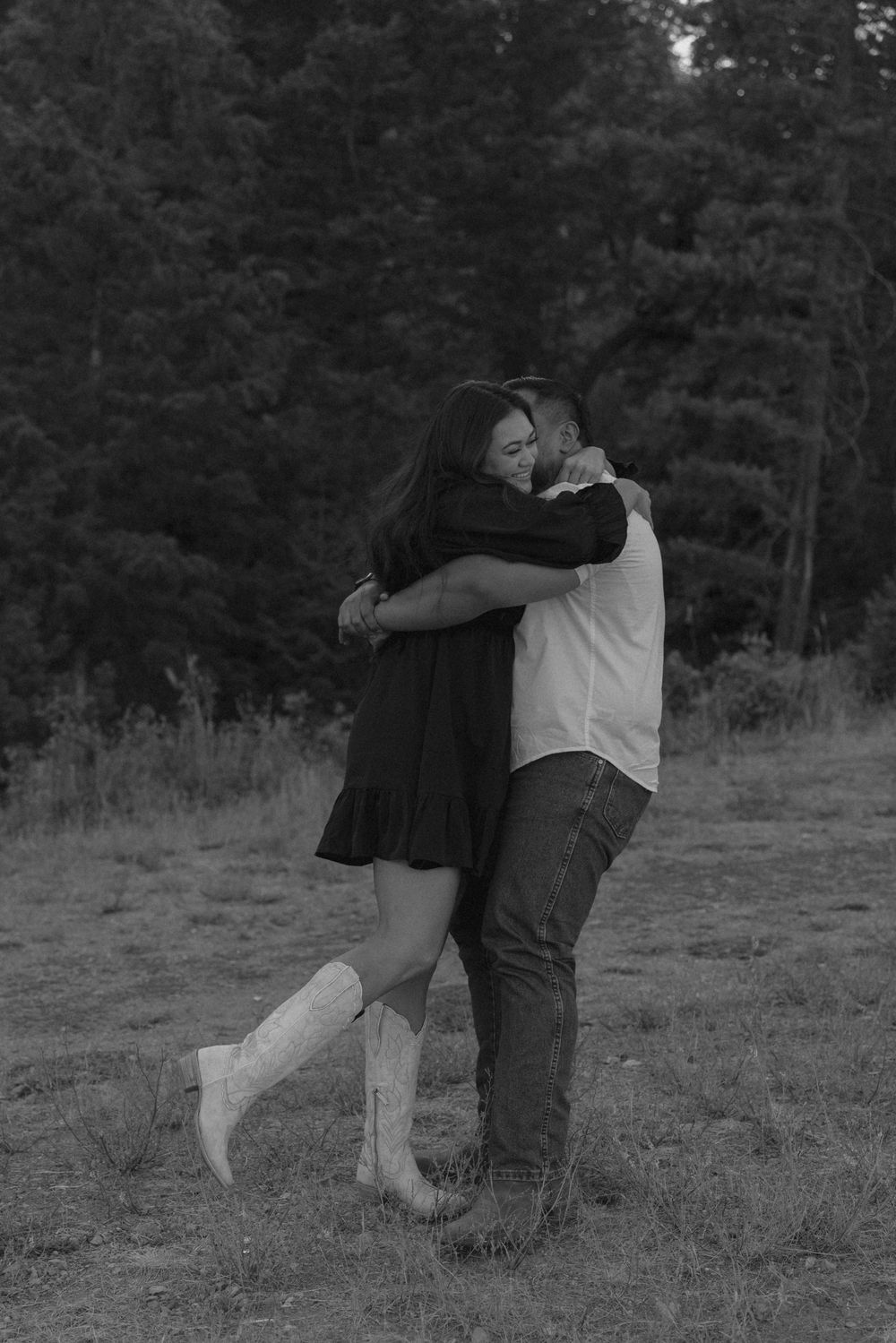 A couple embraces in a black and white photo with forest scenery behind them.