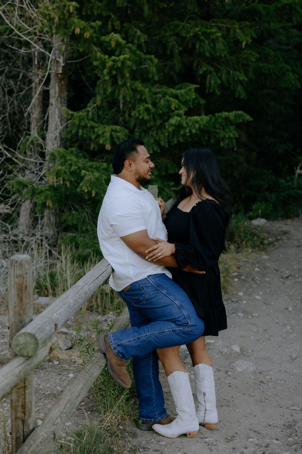 A couple sits together on a rustic wooden fence surrounded by evergreen trees.