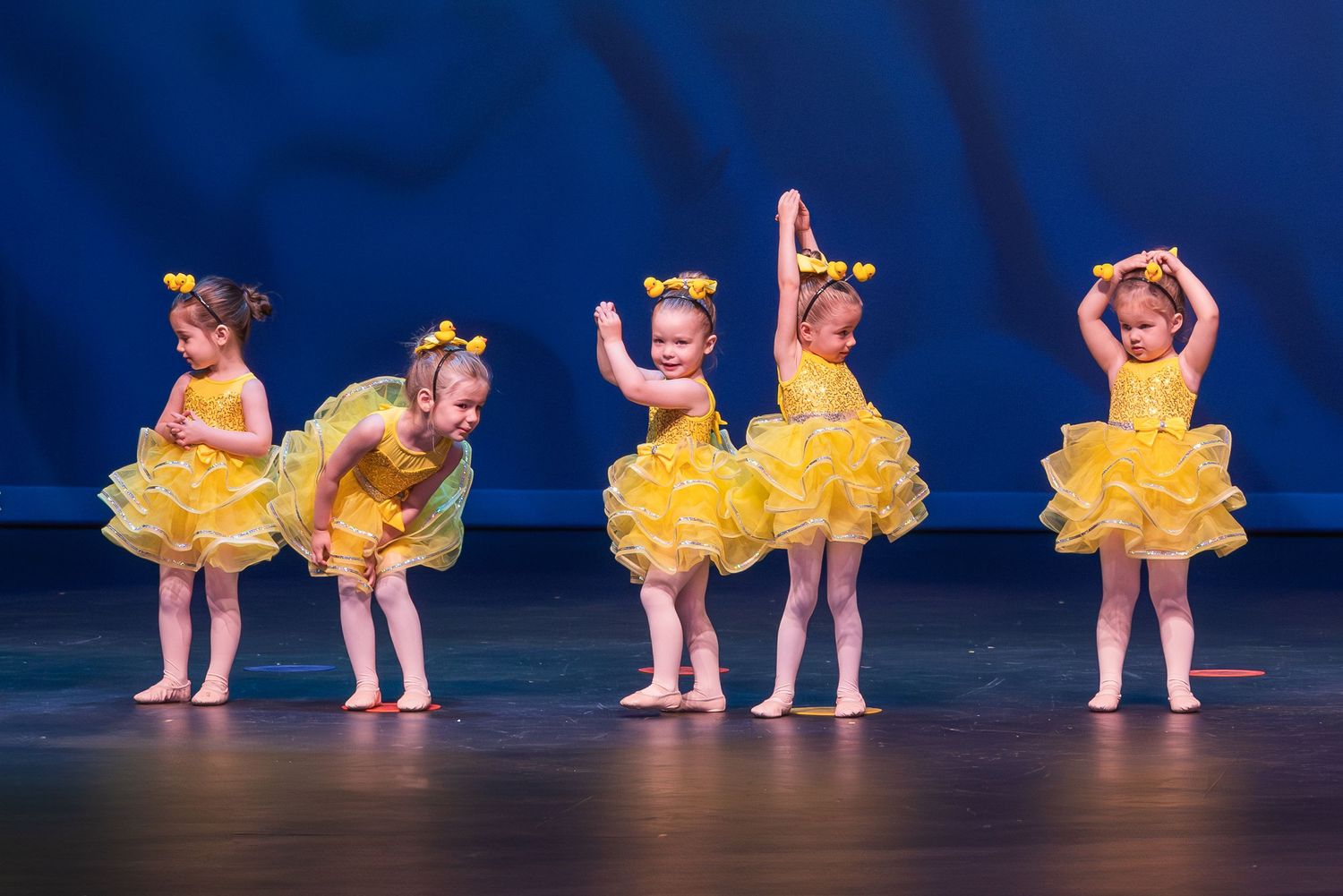 Young ballet dancers in yellow tutus perform a synchronized dance routine on a blue-lit stage.
