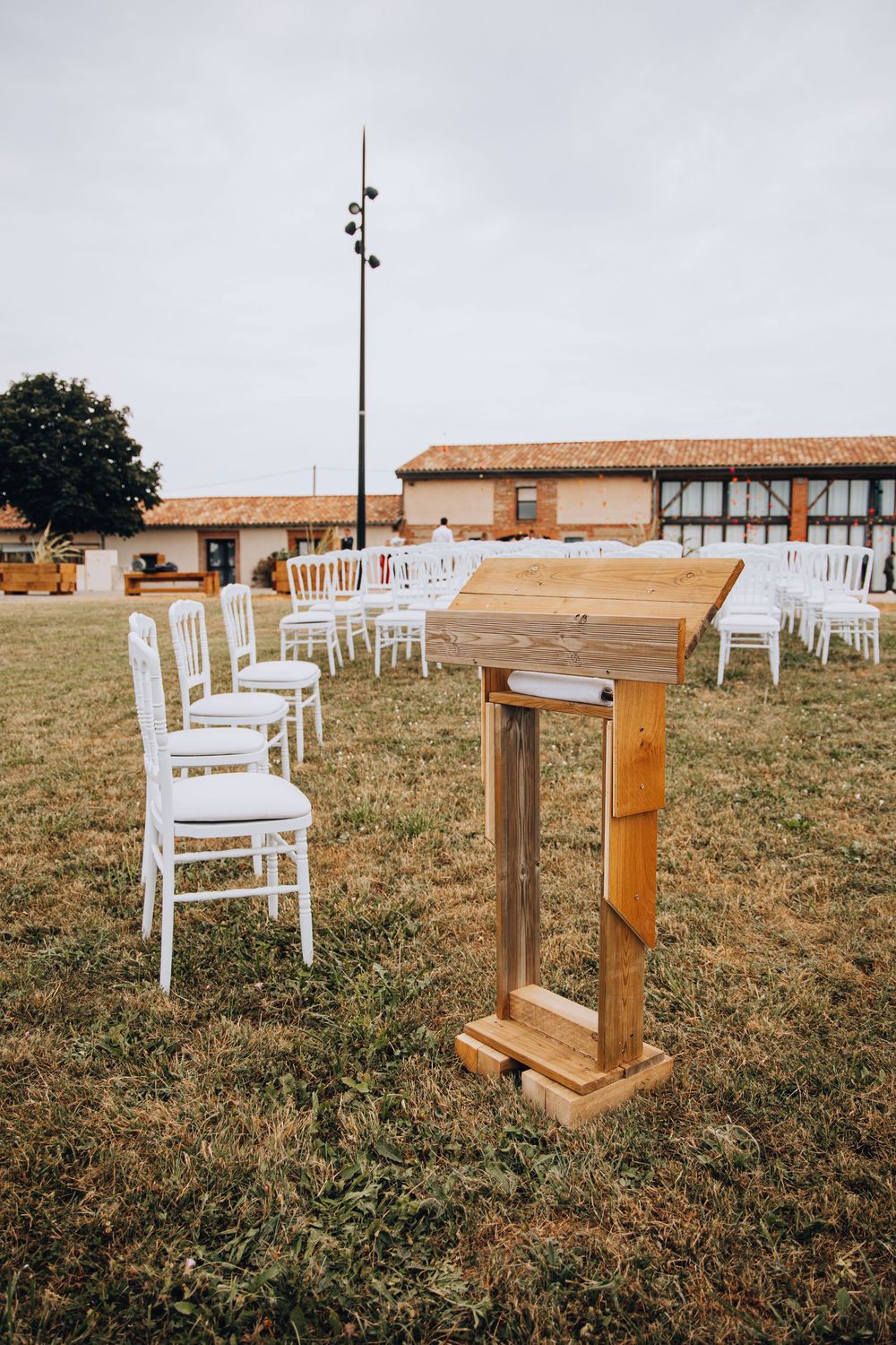 Wooden wedding ceremony podium with white Chiavari chairs arranged on grass outdoors.
