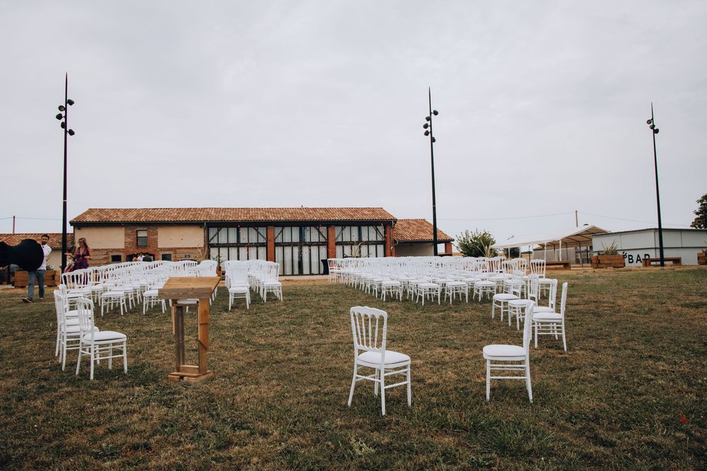 Outdoor wedding ceremony setup with white chairs, wooden table, and a rustic barn backdrop.