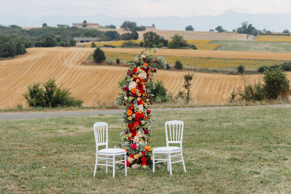 Tall floral column with red and orange blooms flanked by two white chairs overlooking fields.
