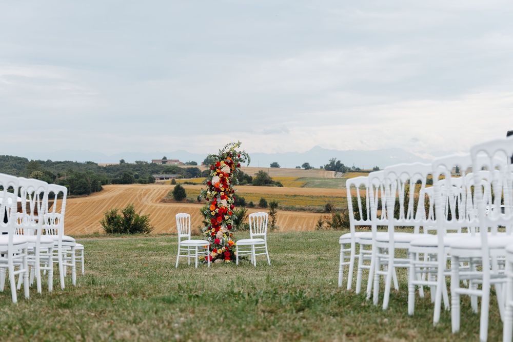 Outdoor wedding ceremony with white chairs, vibrant floral column, and rolling countryside views.