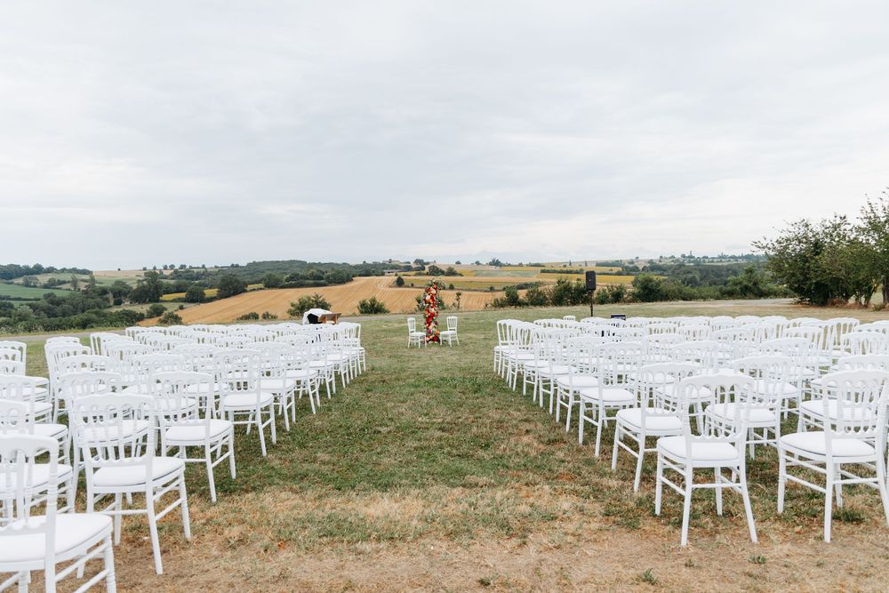 Wide outdoor wedding ceremony setup with white chairs and colorful floral arch amid scenic fields.