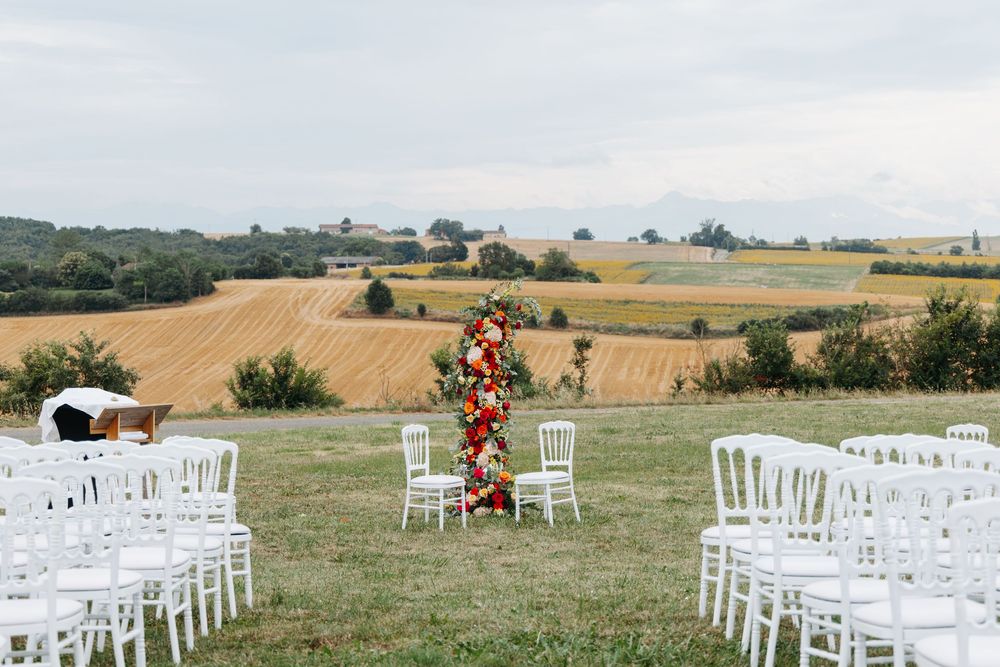 Elegant outdoor wedding ceremony with white chairs, tall floral arrangement, and golden field views.