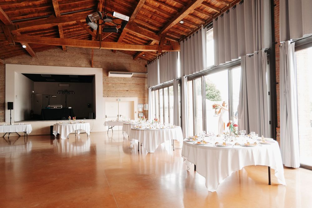 Spacious wedding venue with white draped tables, wooden ceiling, large windows, and a stage area.