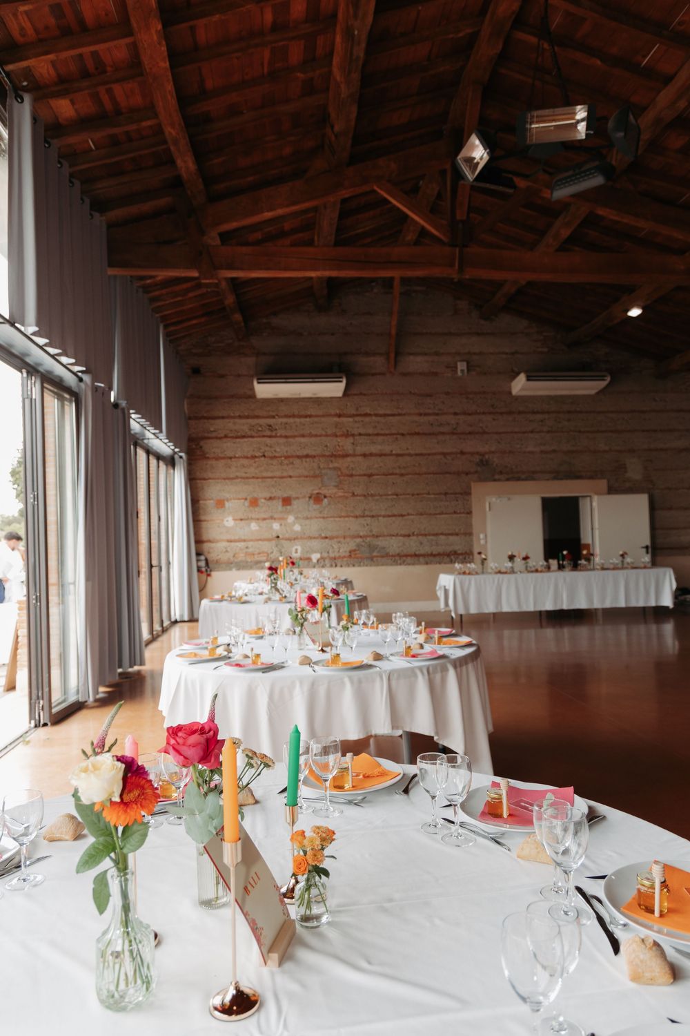 Rustic wedding reception with colorful floral centerpieces, candles, and orange napkins on white tables.
