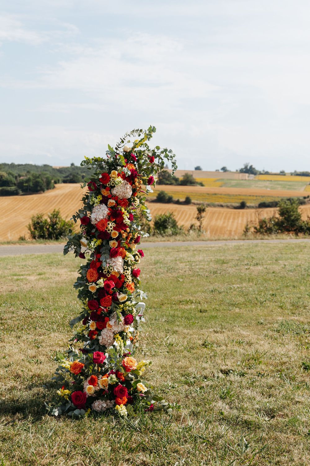 Tall floral column arrangement with red, yellow, and white flowers in an outdoor countryside setting.