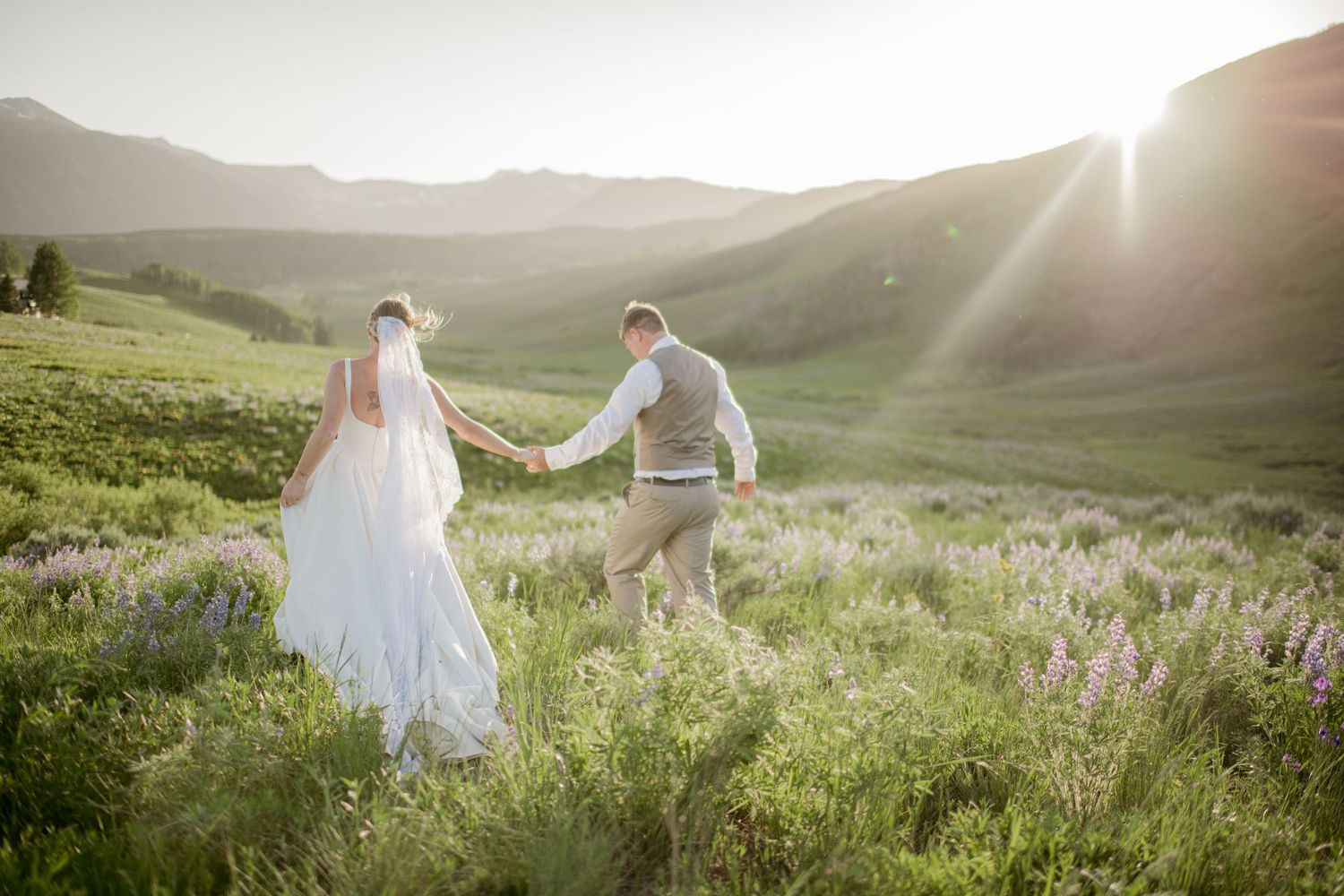 A couple in wedding attire poses together against a scenic mountain landscape with green trees in the background.