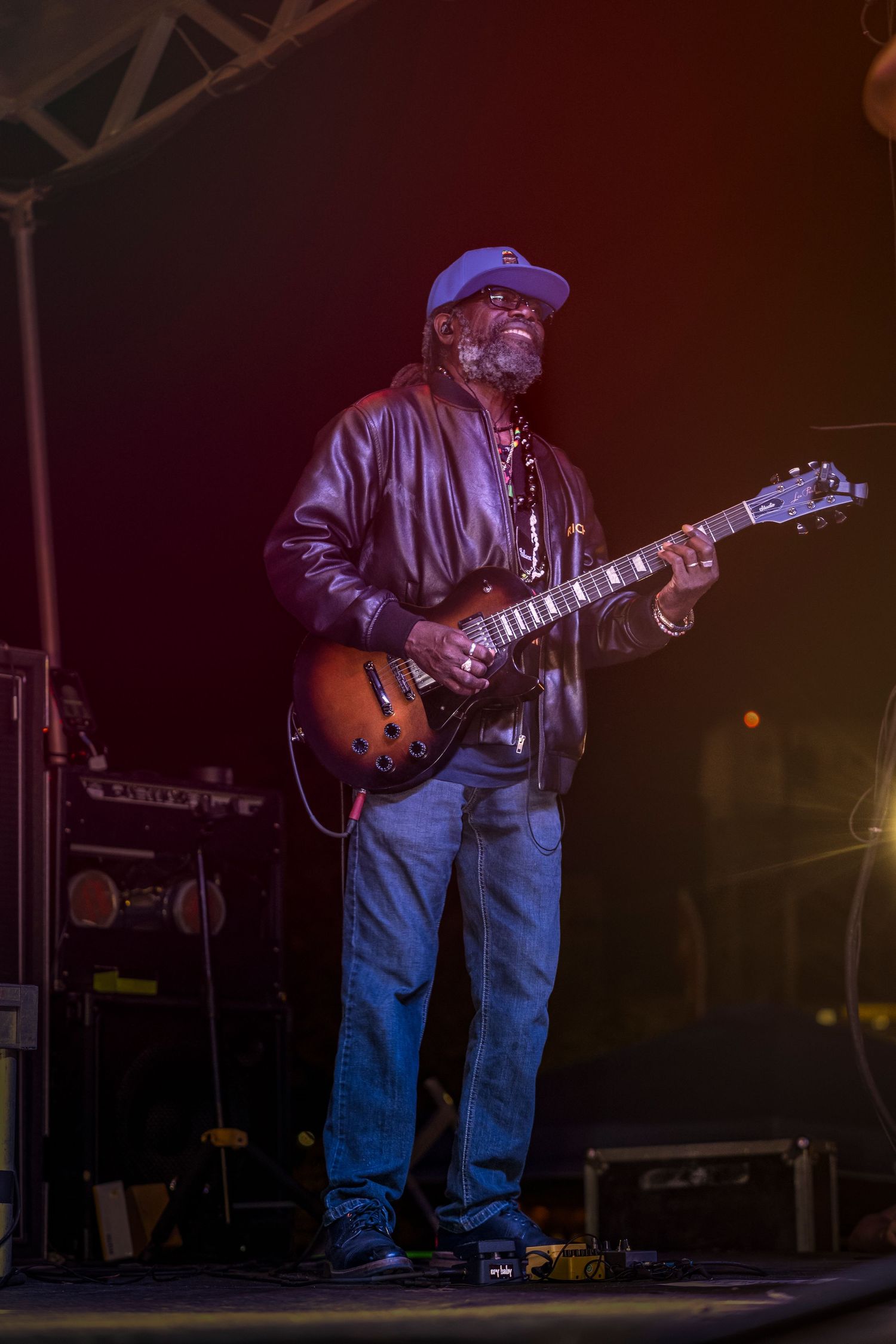 Blues guitarist in denim and cap performs on stage in dramatic lighting during a live concert performance.