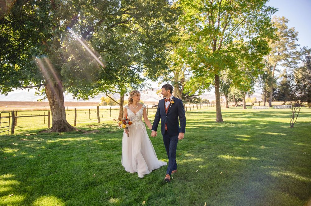 A couple strolls together on lush green grass with tall trees creating a natural outdoor backdrop.