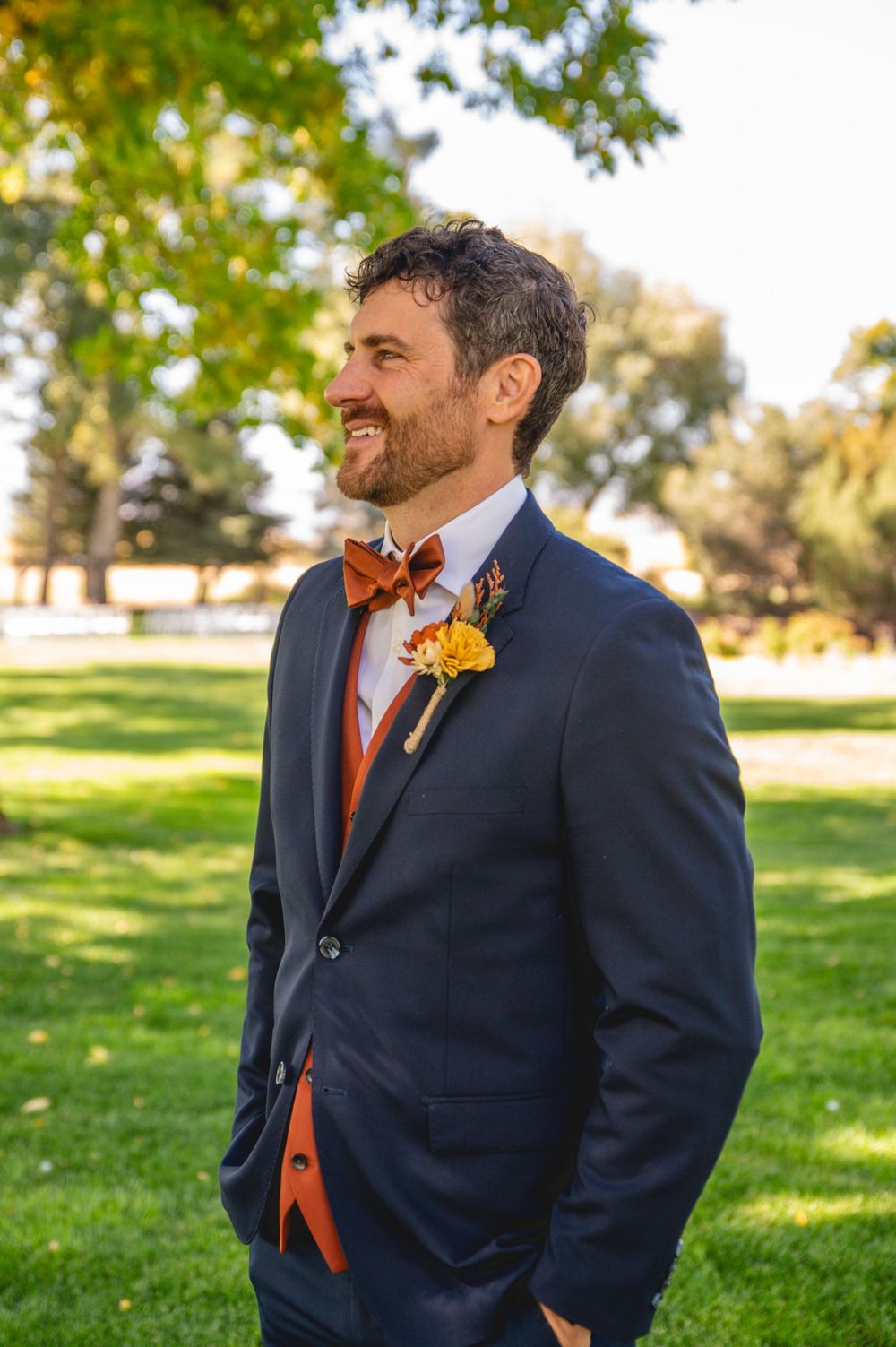 A groom in a navy suit and orange bow tie smiles while standing outdoors on a sunny autumn day.
