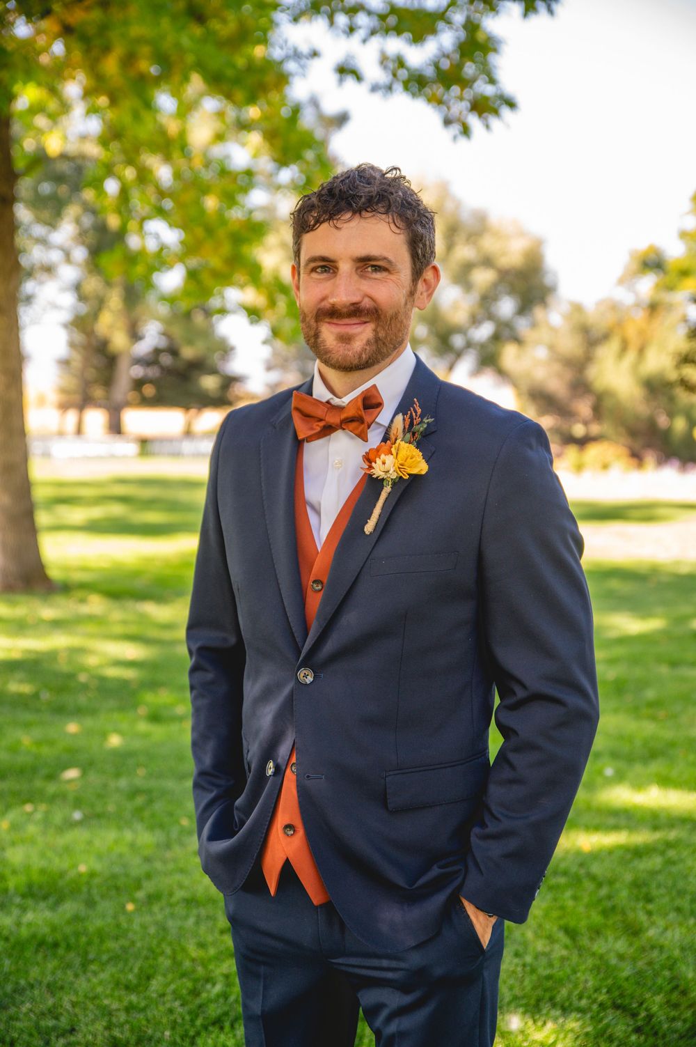 A groomsman in a navy suit with orange vest and bow tie poses outdoors on a sunny autumn day.