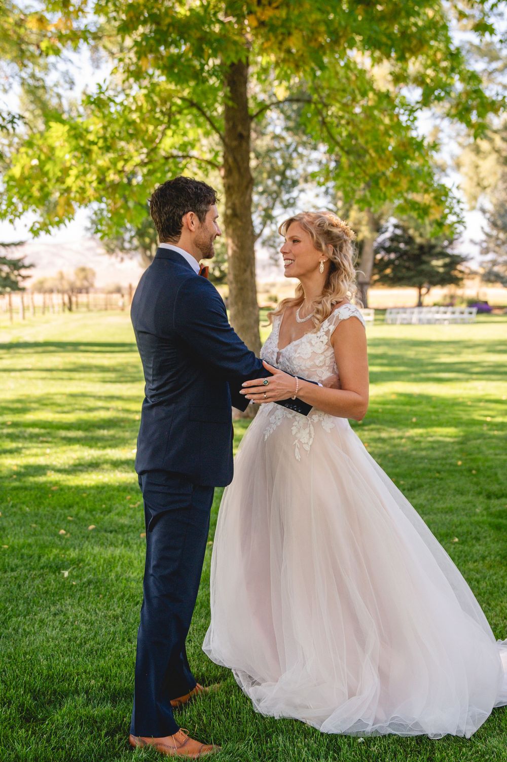 A bride and groom share a joyful moment dancing together on a grassy lawn surrounded by trees on their wedding day.