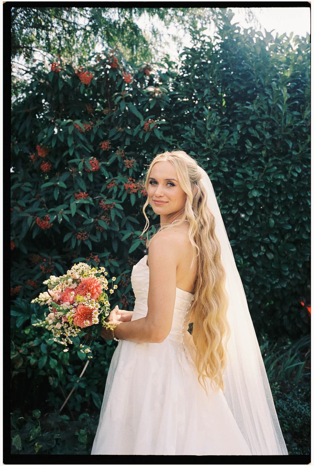 A bride in a flowing white wedding gown poses in front of lush greenery and red flowers in a garden setting.