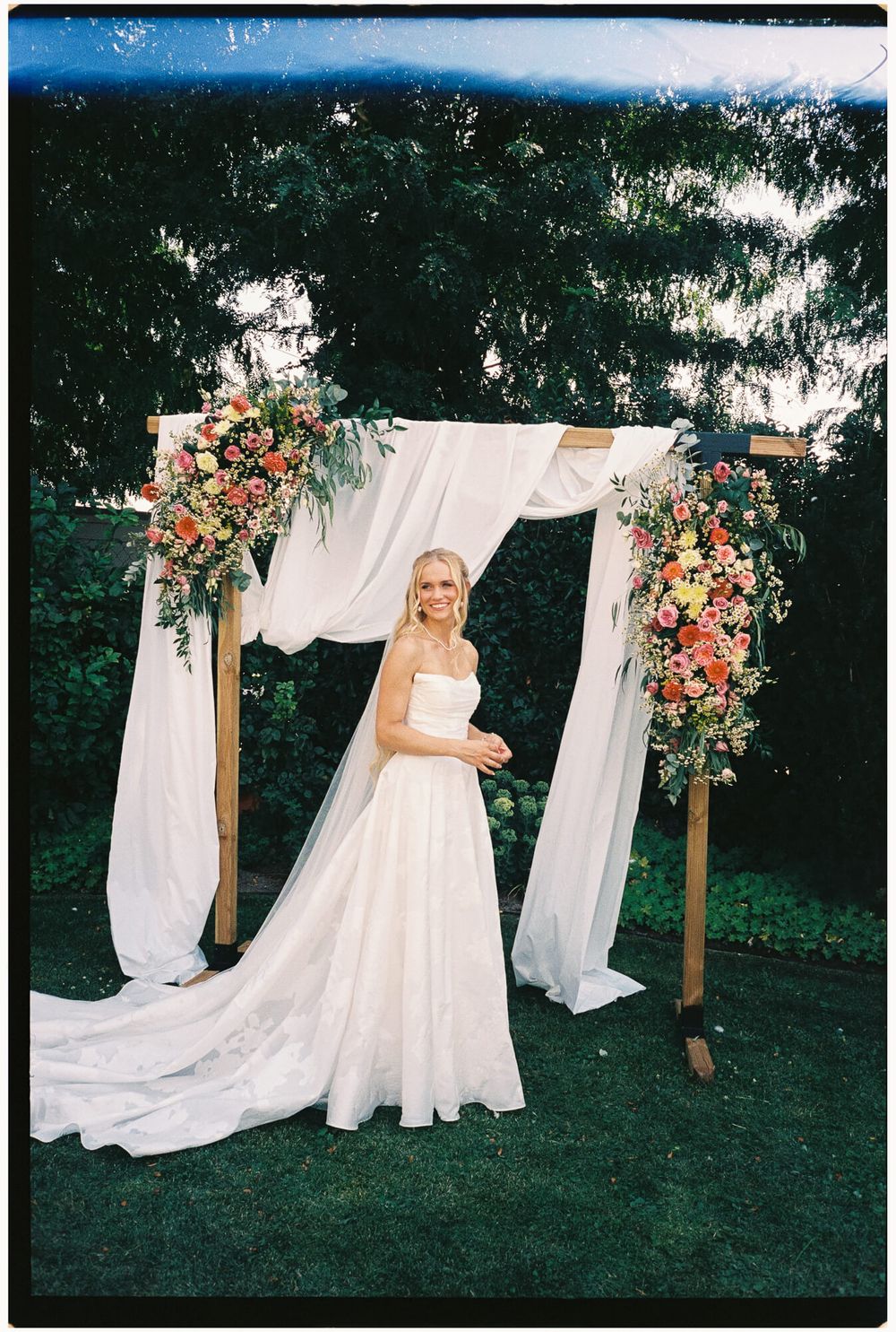 A wedding arch decorated with white fabric and coral flowers stands on a green lawn during an outdoor ceremony.