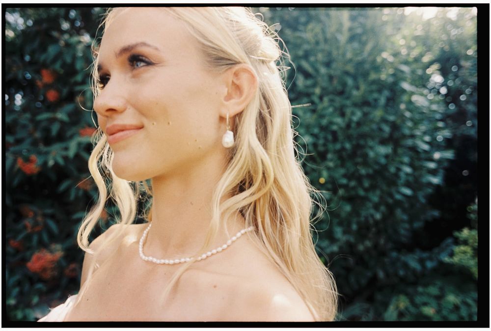 A person with blonde wavy hair wearing pearl jewelry and a pink dress poses outdoors against green foliage.