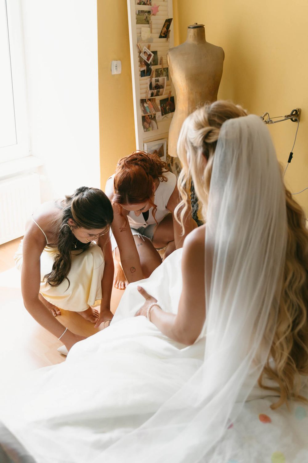 A group gathers around a bride in her white wedding dress and veil during wedding preparations.