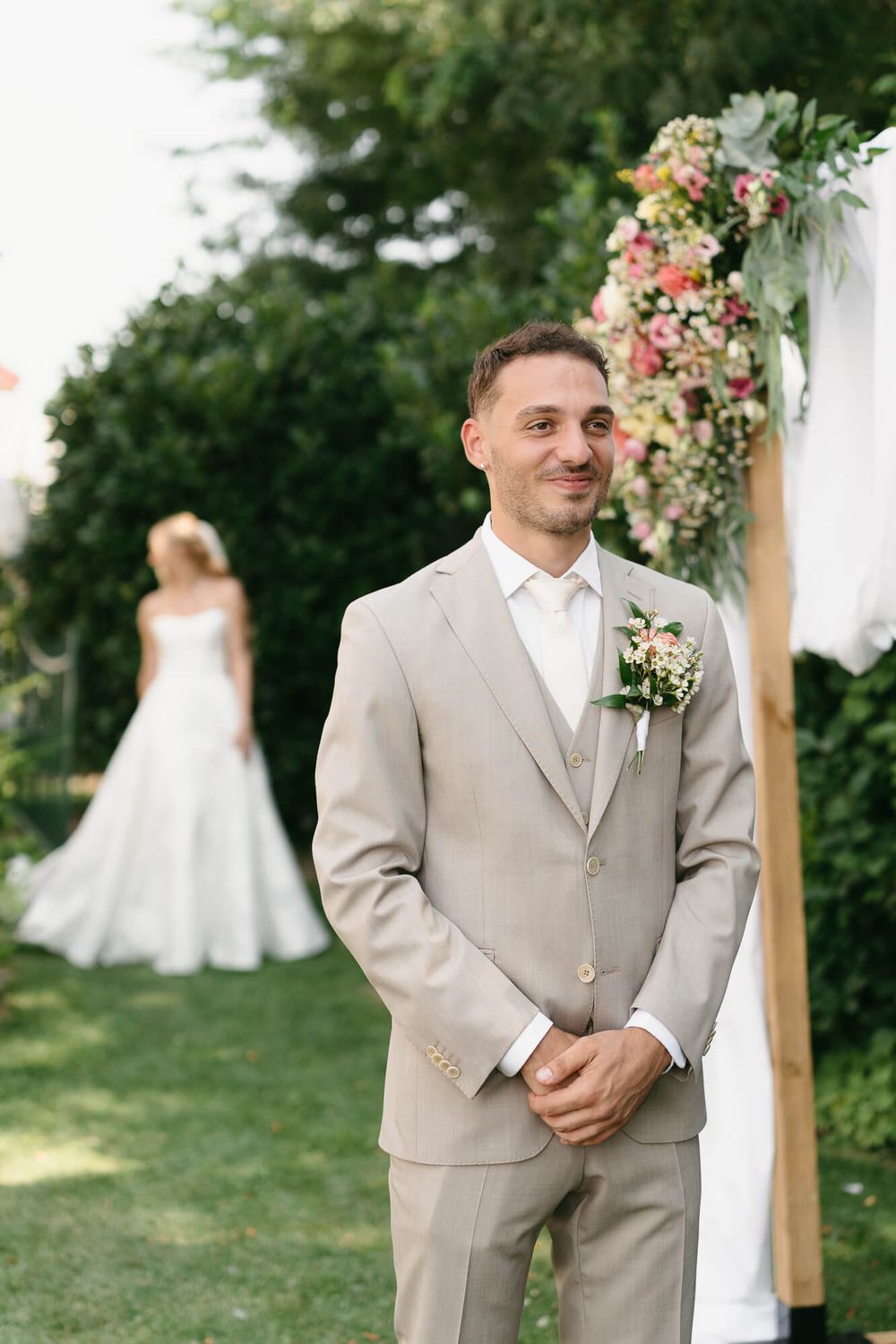 A groom in a light beige suit stands in a garden setting with floral wedding arch decorations.