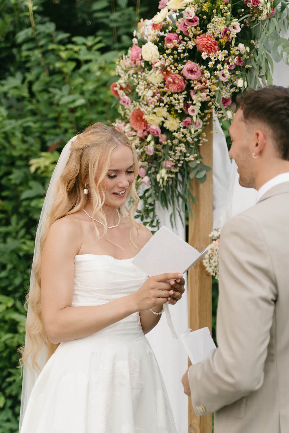 A couple exchanges rings during an outdoor wedding ceremony beneath a floral arch decorated with pink blooms.