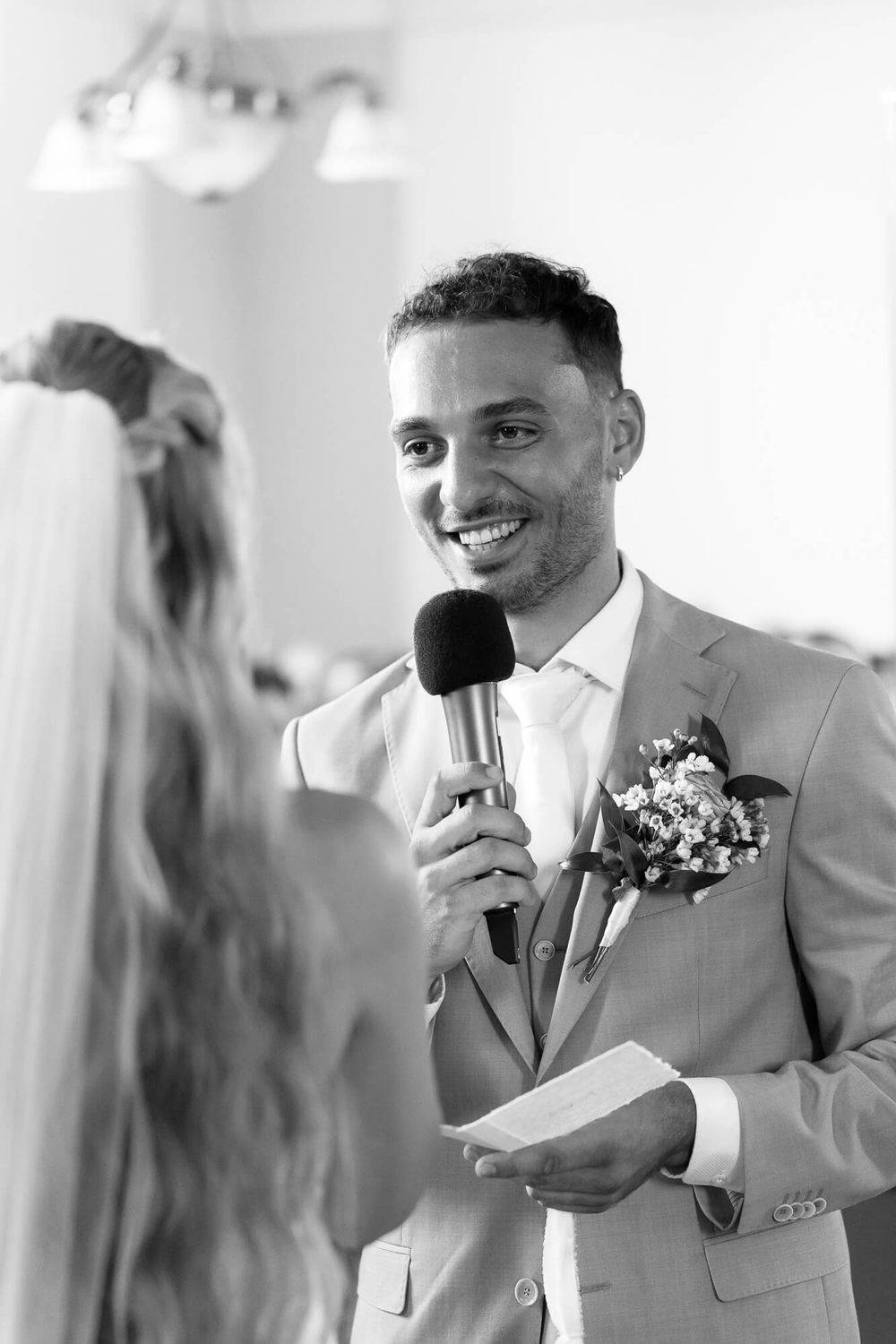 A smiling groomsman in a light gray suit holds a microphone during a wedding reception speech.