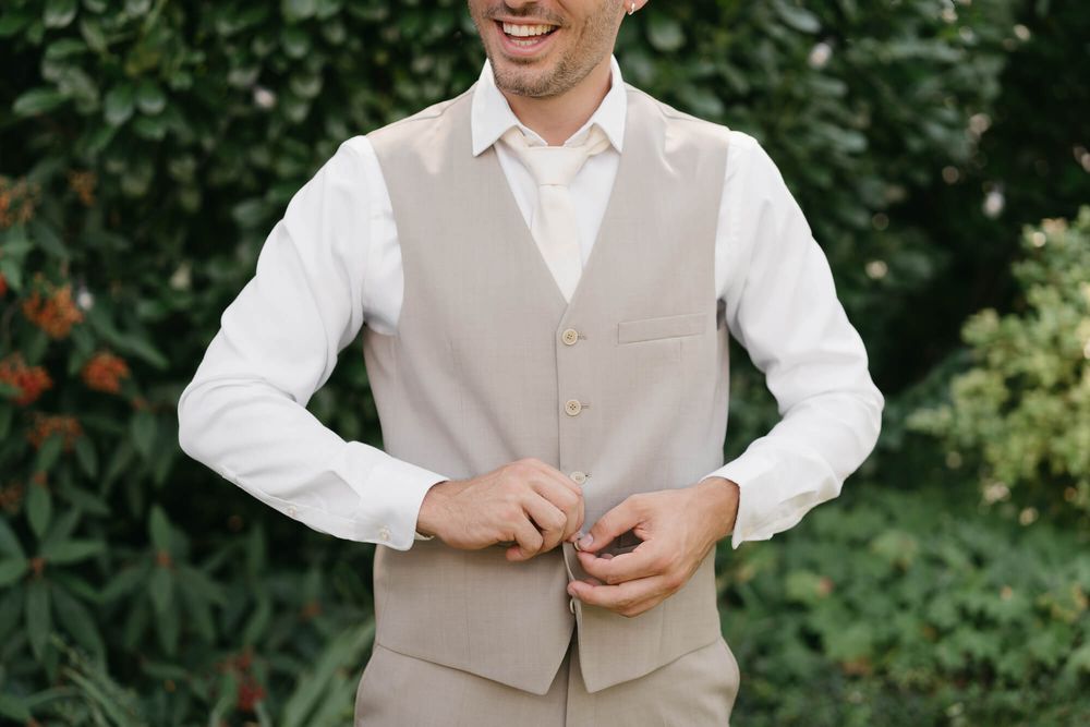 A person in a light beige vest and white dress shirt stands outdoors against a lush green garden background.