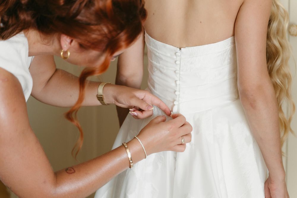 A person helps fasten the buttons on a white wedding dress corset back during wedding preparations.