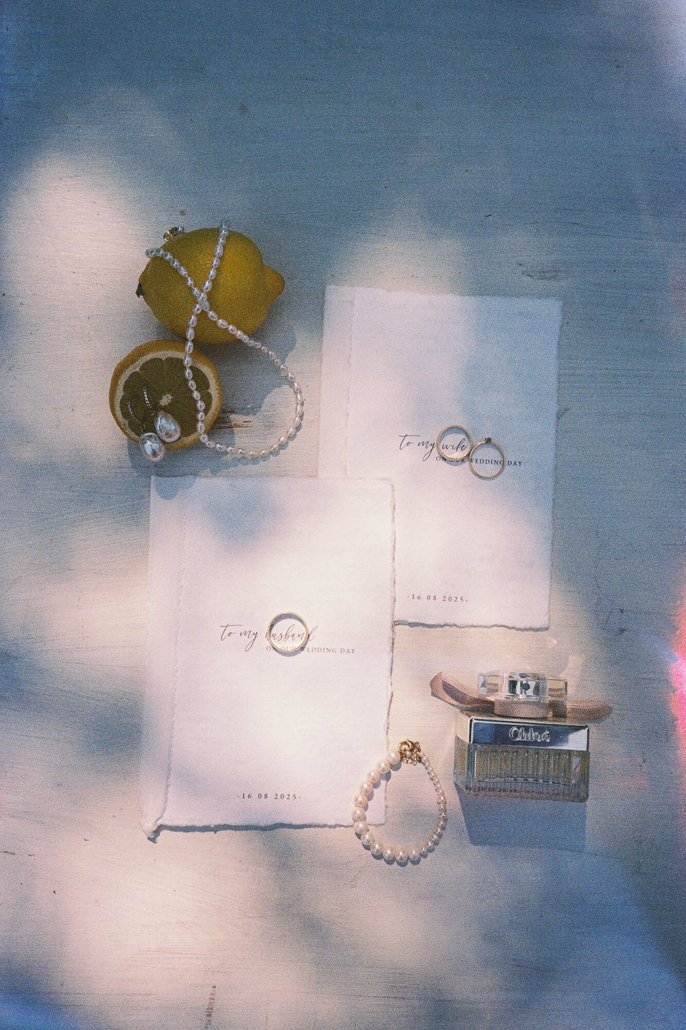 Flat lay of wedding stationery with pearl jewelry, Chloe perfume, and lemon slices on a soft blue background.