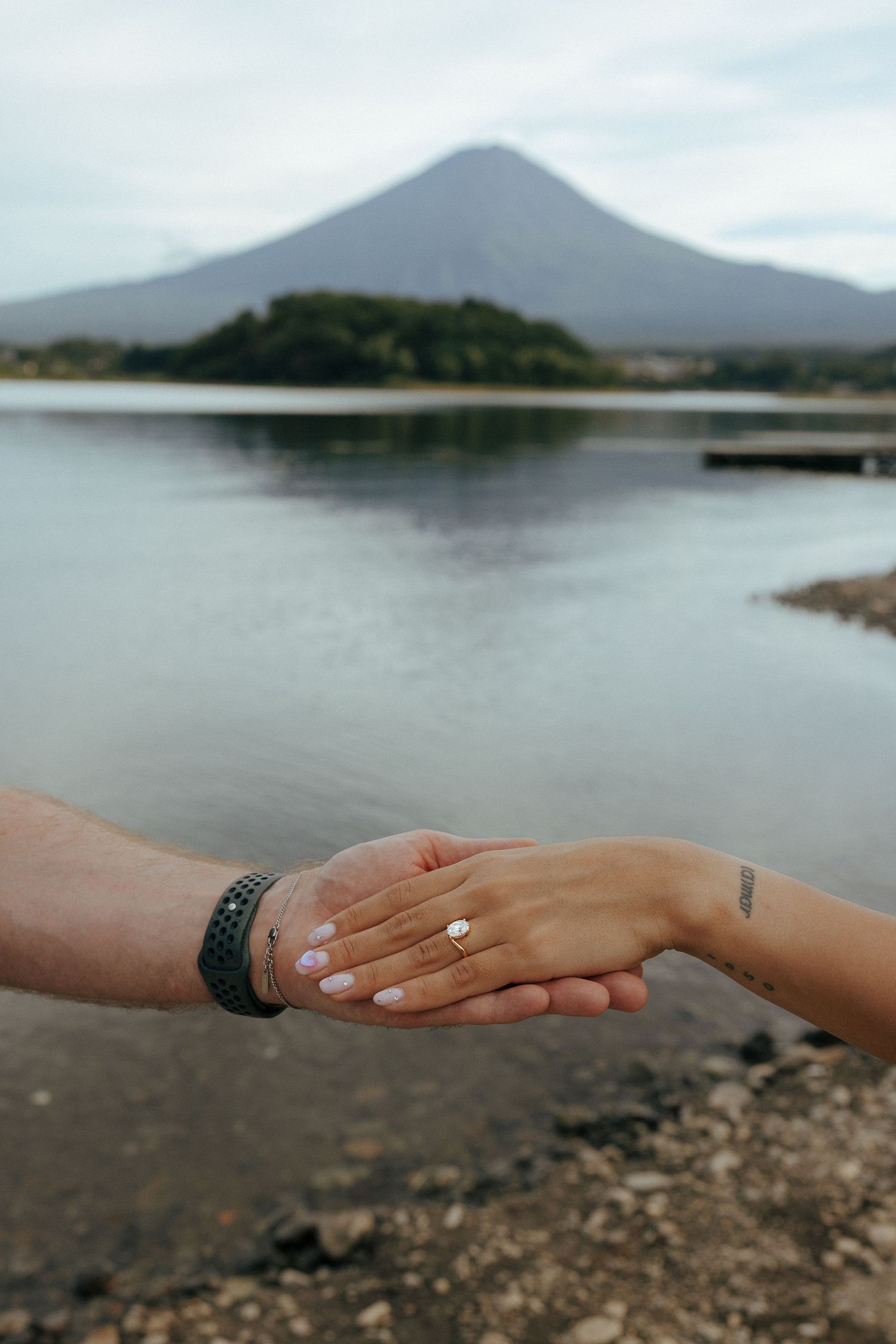 Two hands reach out across a rocky lakeshore with Mount Fuji and water in the misty background.