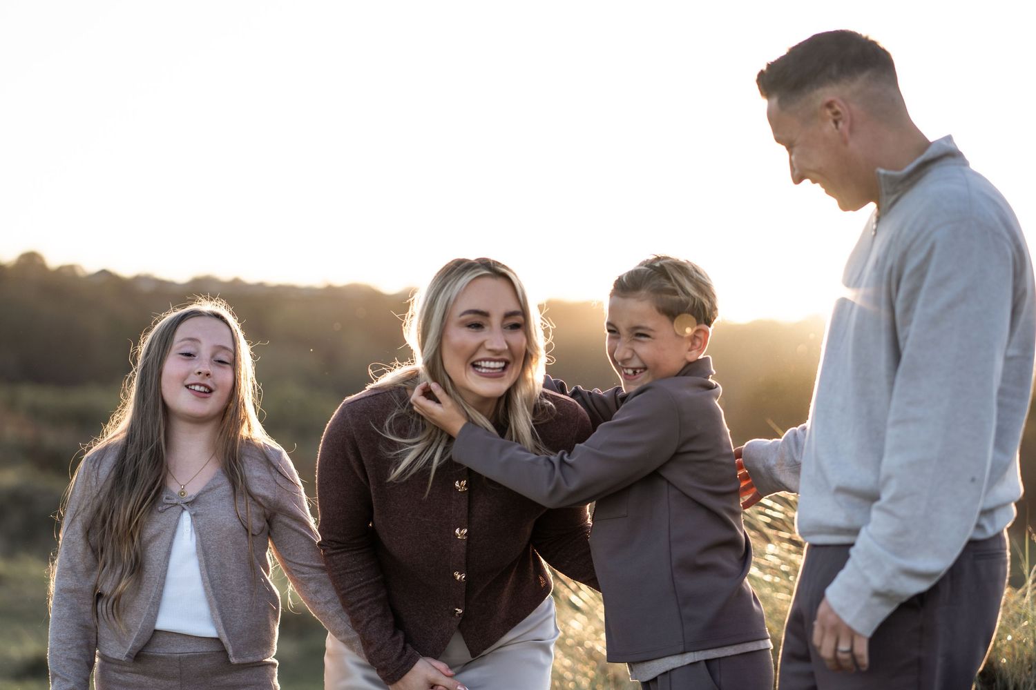 A group of people laugh and embrace while standing together in a field during golden hour sunset.