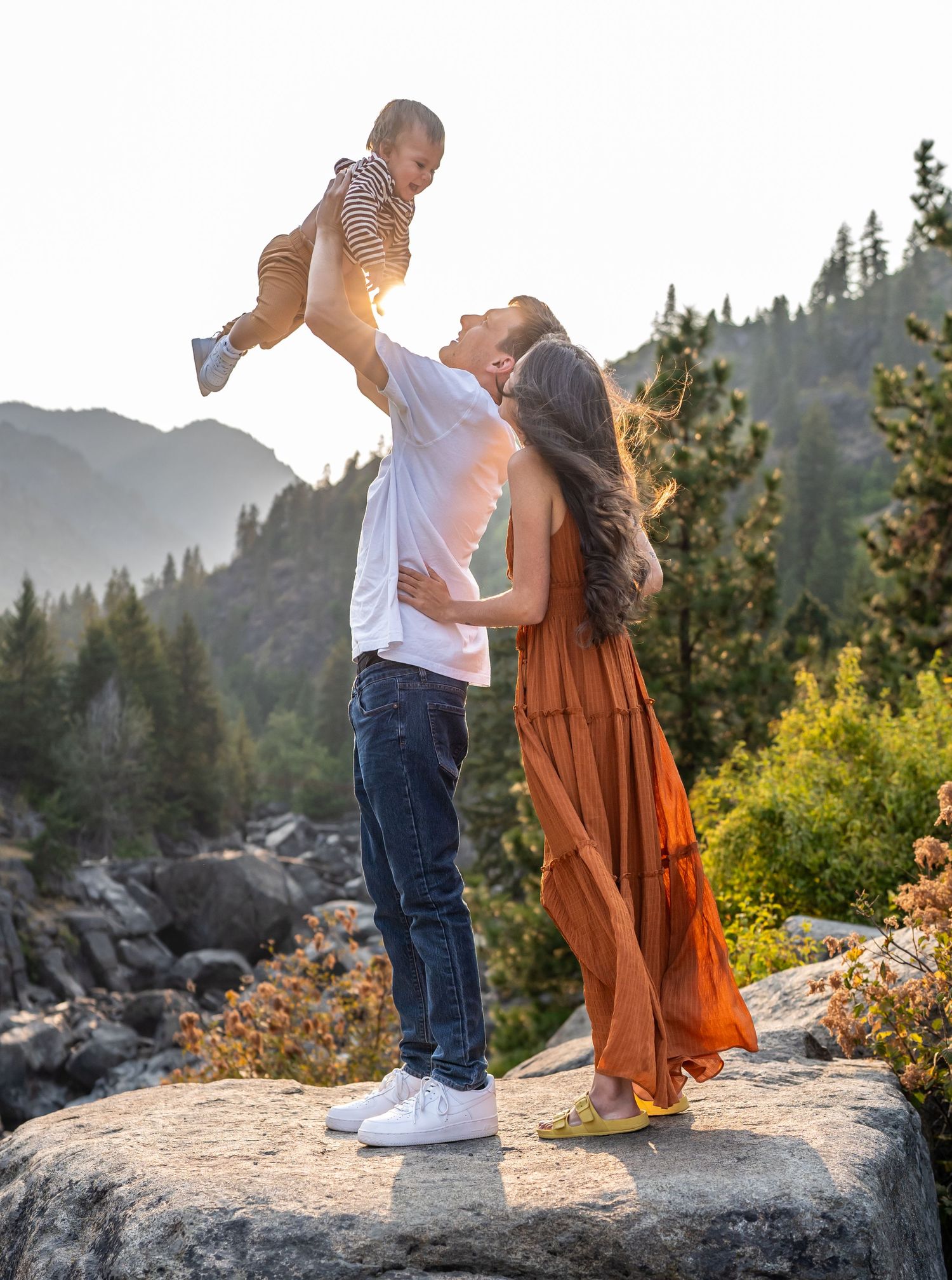 Parents playing with their baby on a scenic mountain overlook at golden hour.