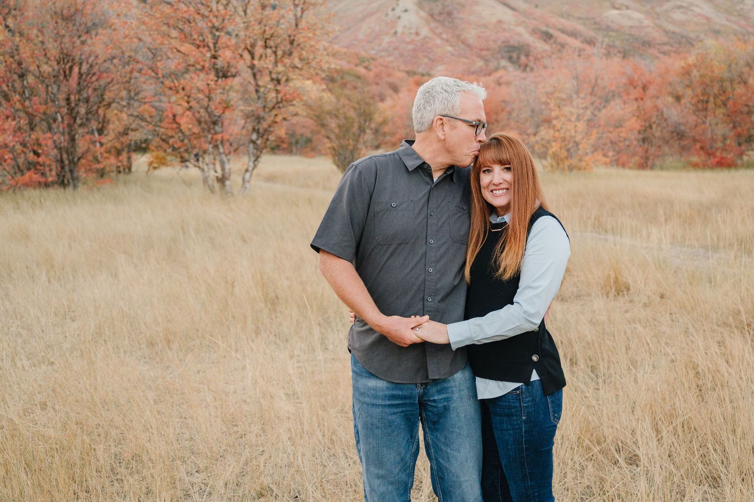 A couple embraces while standing in a golden autumn field with reddish hills and bare trees in the background.