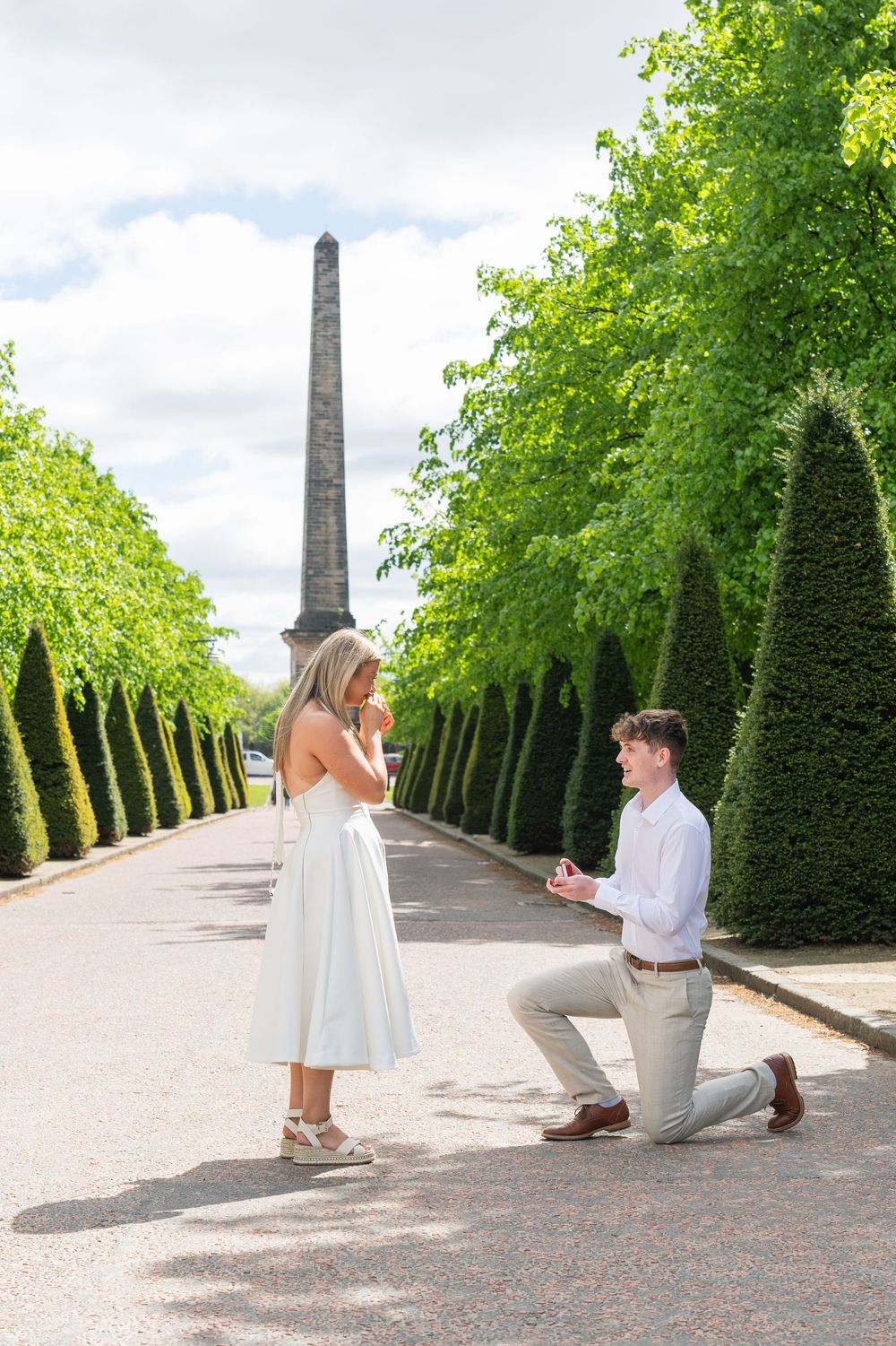A romantic marriage proposal takes place in a beautiful garden path lined with manicured trees and obelisk monuments.