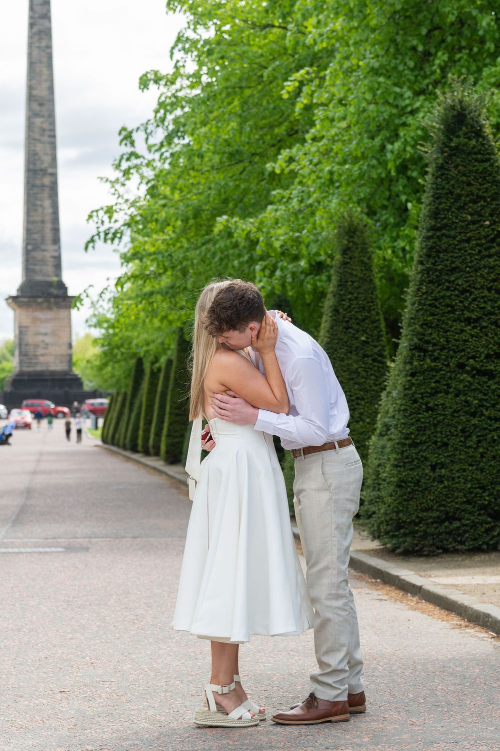A couple shares an intimate moment on a tree-lined pathway with tall hedges and stone monuments in the background.