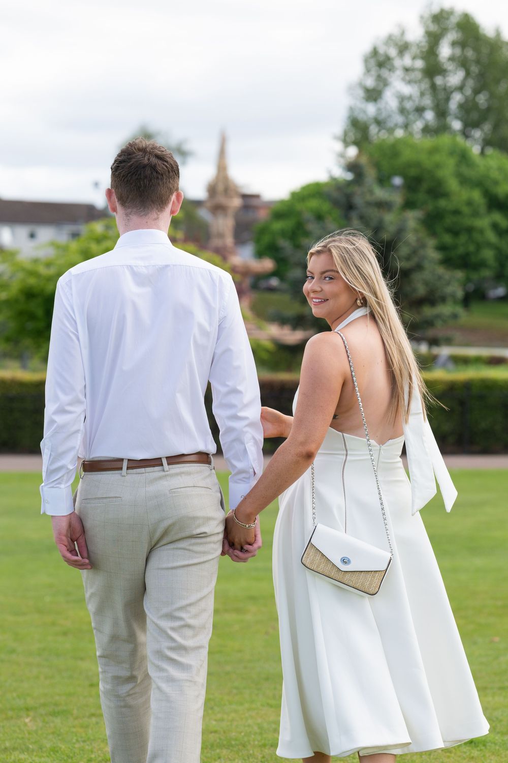 A couple in white and beige formal attire walk hand in hand across a green lawn on a sunny summer day.