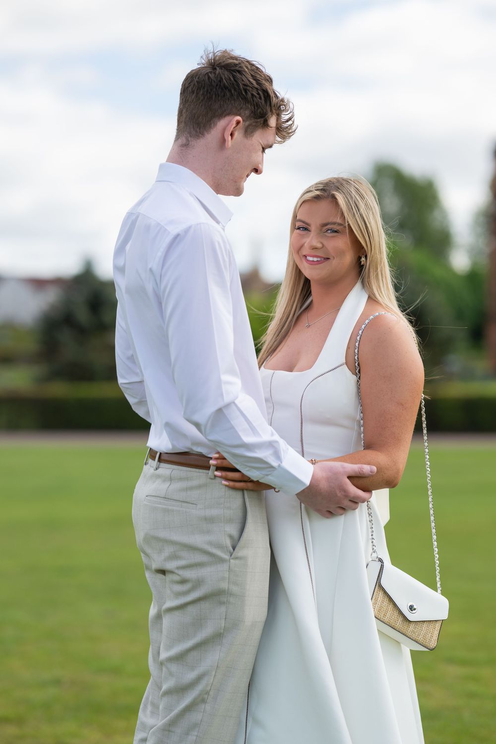 A couple shares an intimate moment in formal white attire while standing together in a grassy outdoor setting.
