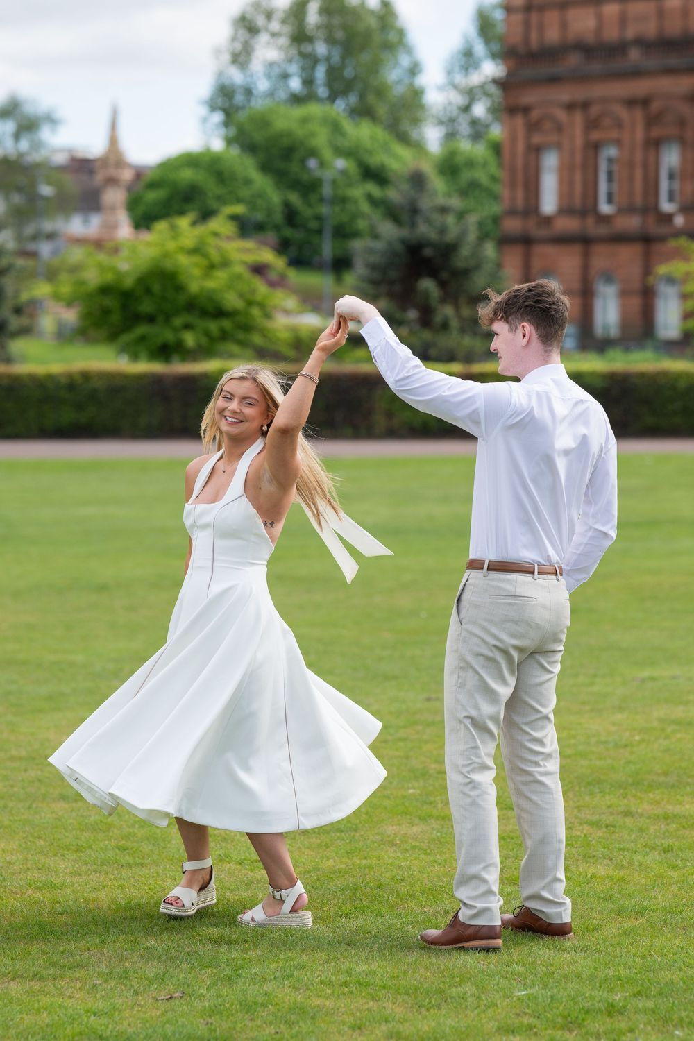 A couple dances playfully on a green lawn in front of a brick building, with the woman twirling in a white dress.