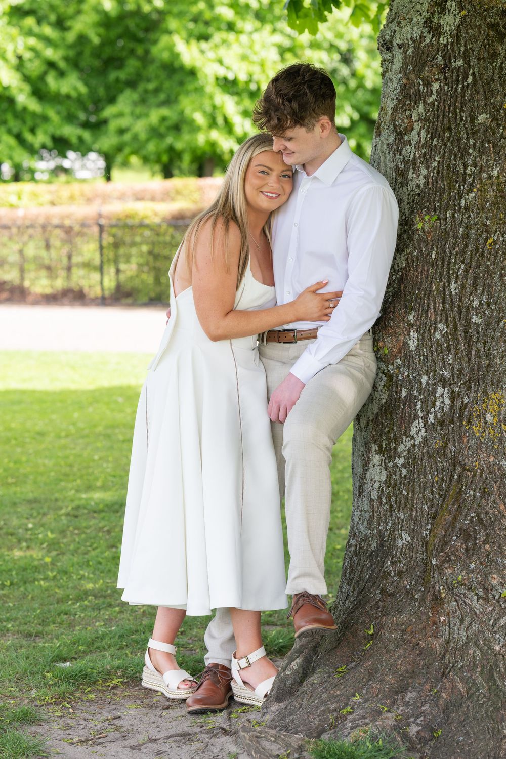 Romantic couple in white clothing embraces against a large tree trunk in a sunny outdoor park setting.