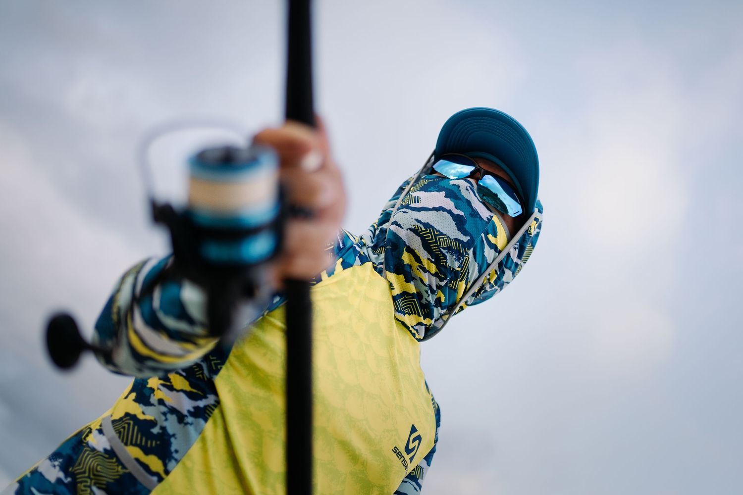 Close-up of hands holding a fishing rod with blue and yellow camouflage sleeves against cloudy sky.