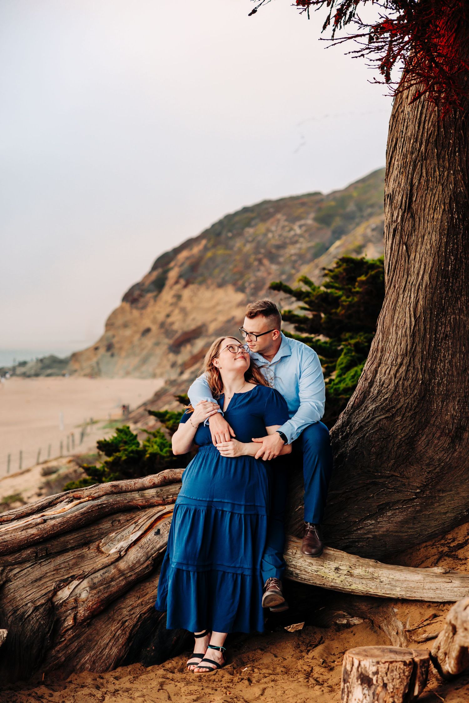 A couple in blue attire share an intimate moment while sitting on driftwood at a scenic coastal beach.