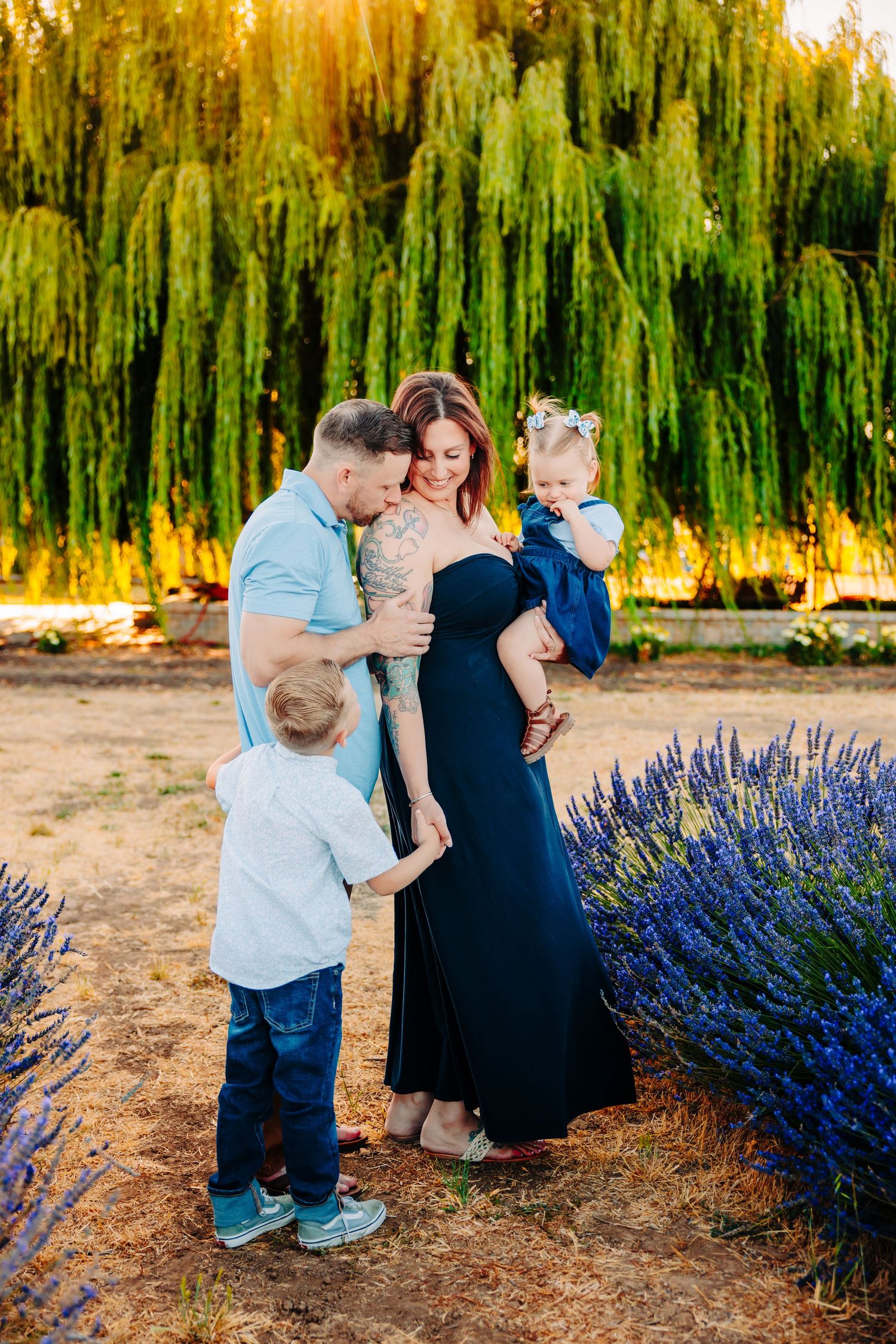 A family shares tender moments by a vibrant lavender garden at sunset with weeping willows in the background.