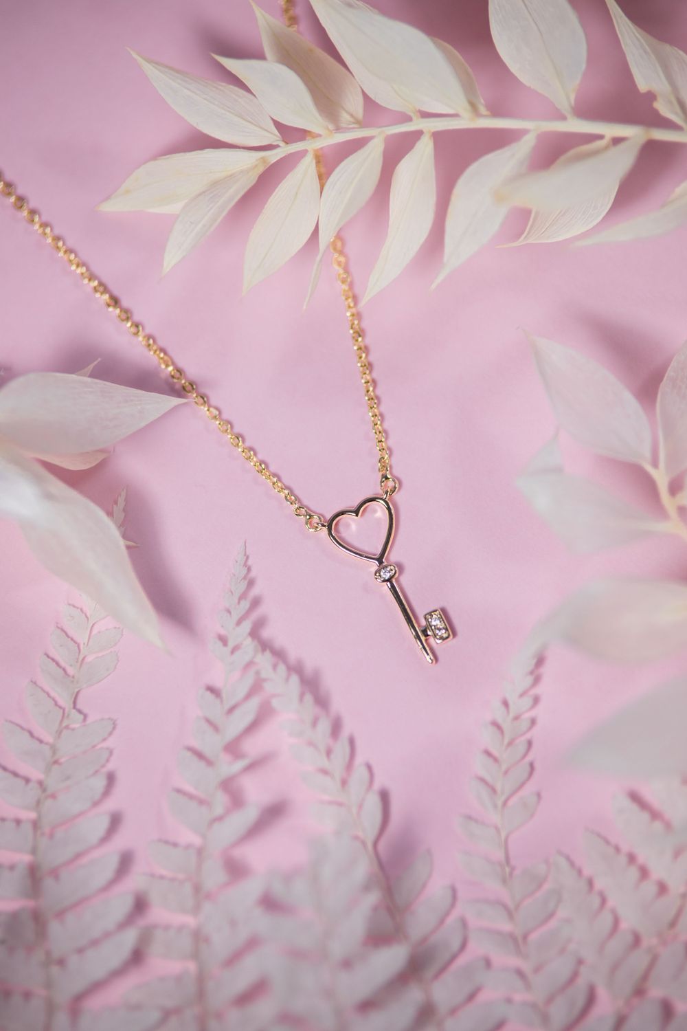 Delicate gold key necklace with heart design against pink background with white leaves.