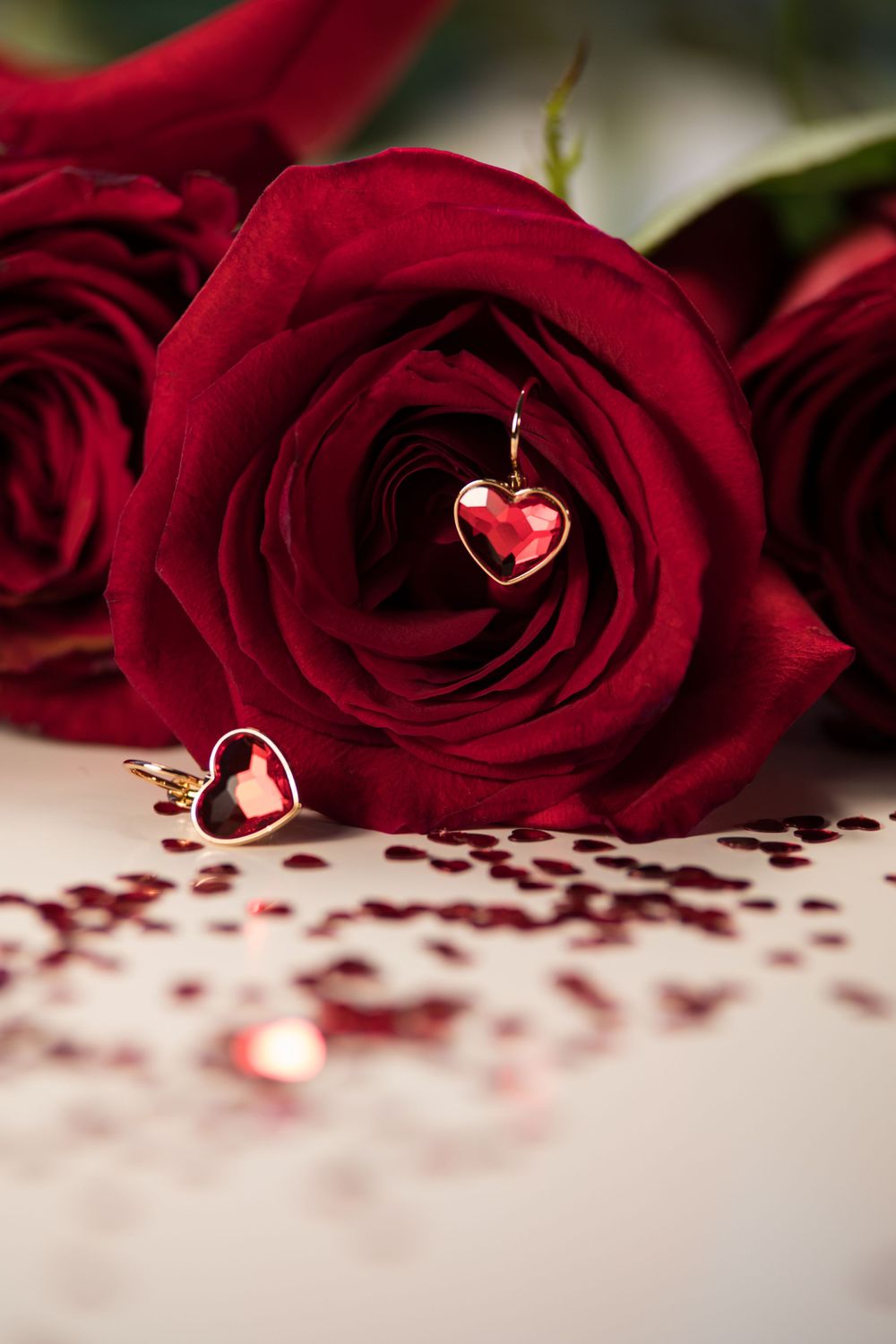 Red heart pendant nestled in deep red rose with scattered rose petals.