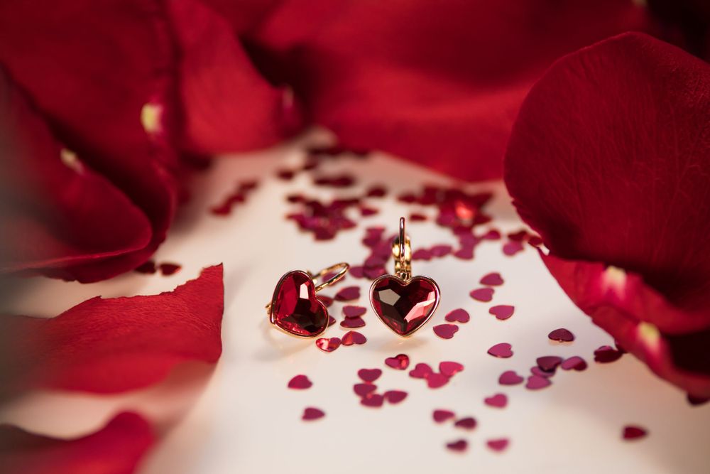 Small heart-shaped jewelry piece surrounded by scattered red rose petals on white surface.