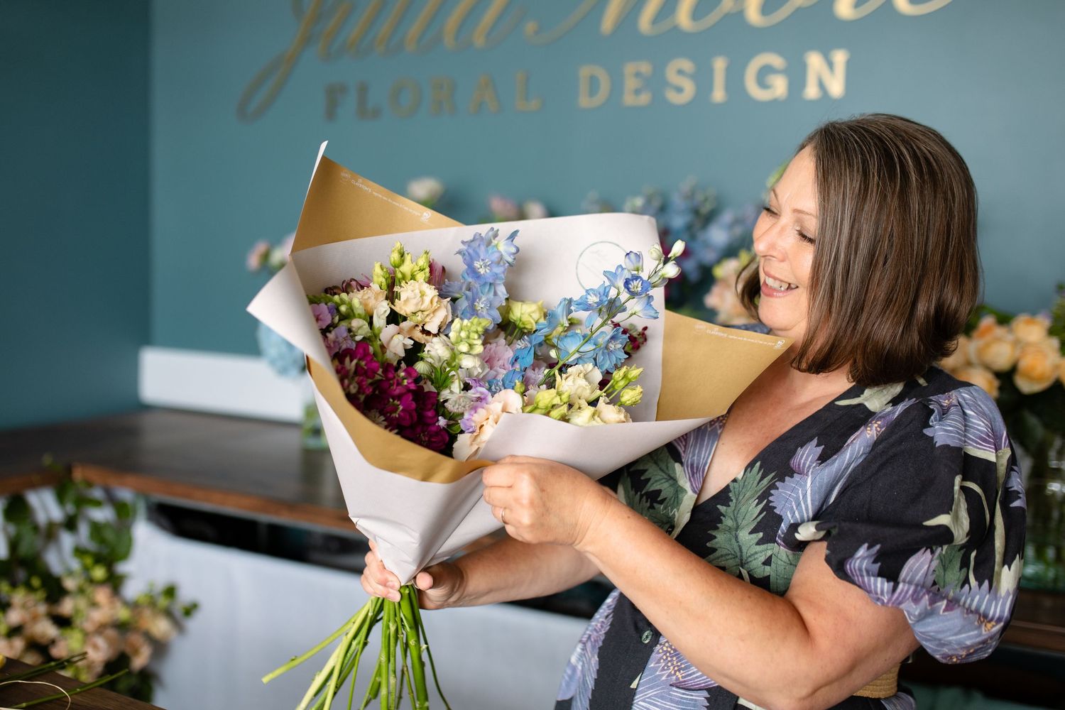 A florist in a floral print dress arranges a vibrant bouquet with blue and purple flowers at a design studio.