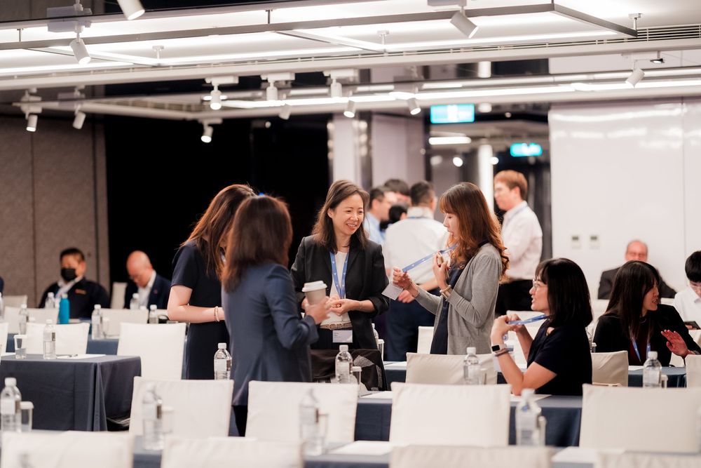 People working at computer stations in a modern office space with white desks and bright lighting.