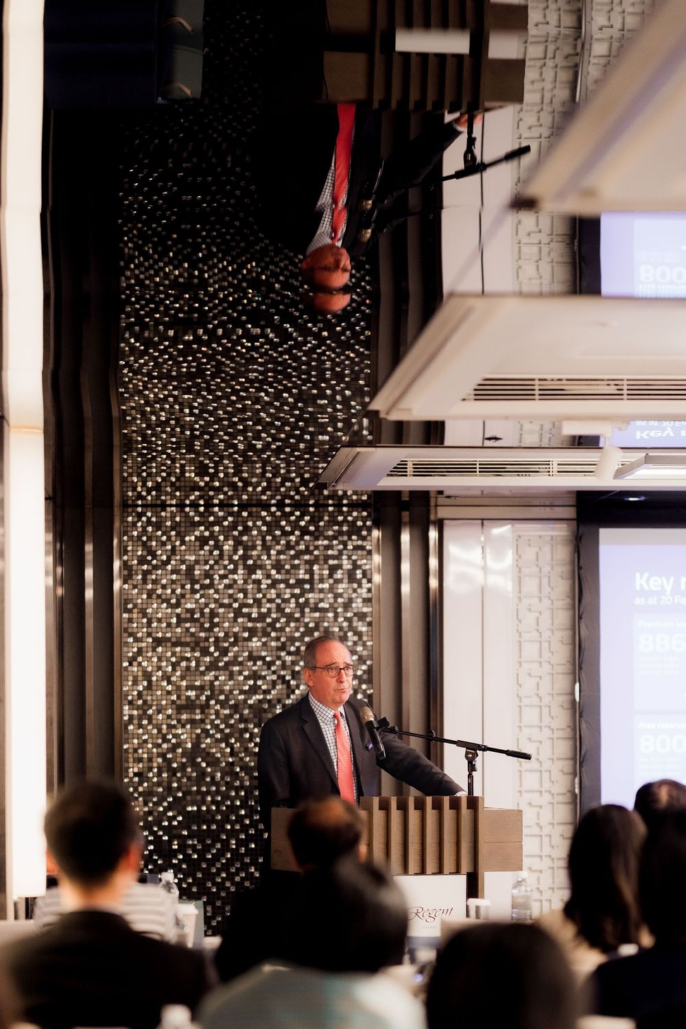 Speaker stands at podium in front of modern geometric wall design during business presentation.