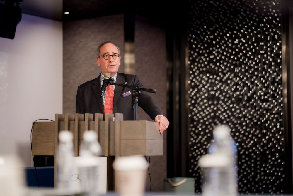 Speaker addresses audience from wooden podium during formal event with decorative lighting elements in background.